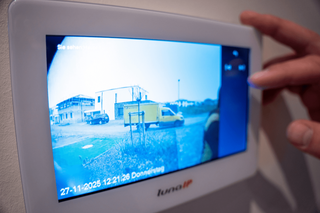 Nahaufnahme eines modernen Innenmonitors einer Video-Türsprechanlage. Auf dem Display ist das Livebild des Eingangsbereichs mit Einfahrt und abgestelltem Fahrzeug zu sehen. Eine Person bedient den Touchscreen, um die Türkommunikation und Zutrittskontrolle zu steuern. Die Aufnahme verdeutlicht die Möglichkeiten moderner Sicherheits- und Smart-Home-Technik.